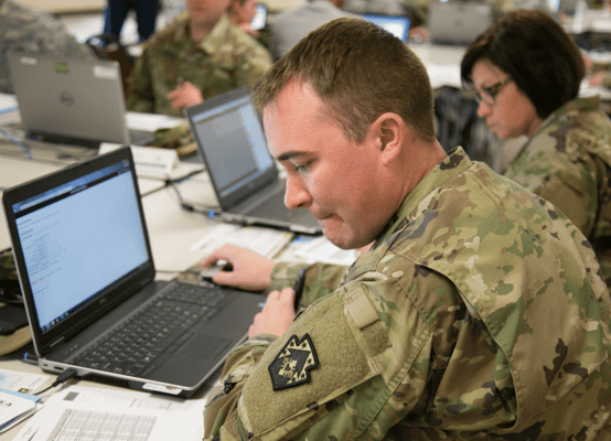 A male military personnel in camouflage uniform working on a laptop at a desk among other soldiers focused on their computers.