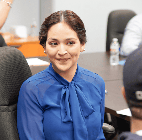 A woman in a blue blouse smiling, seated in a meeting room with blurred figures in the background.